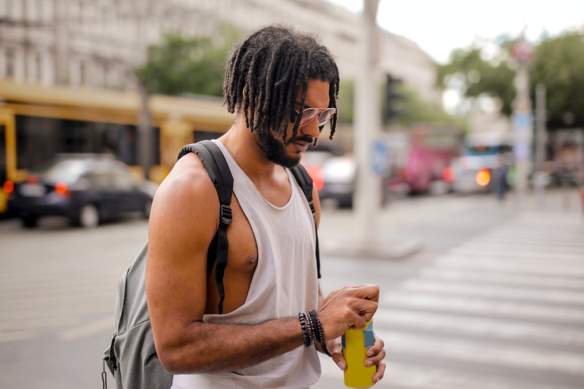 cool ethnic man with dreadlocks drinking beverage while walking along street in city