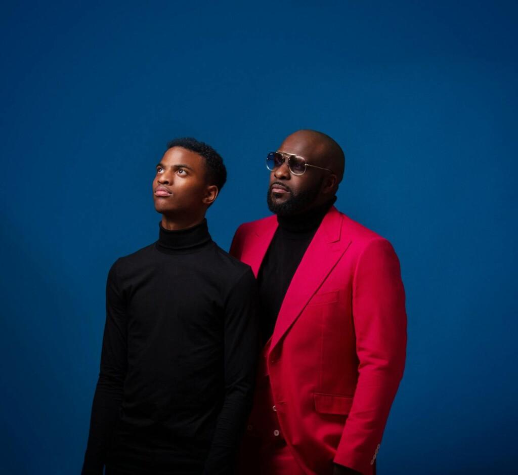 Posed portrait of a father and son in contrasting outfits with a blue studio background.