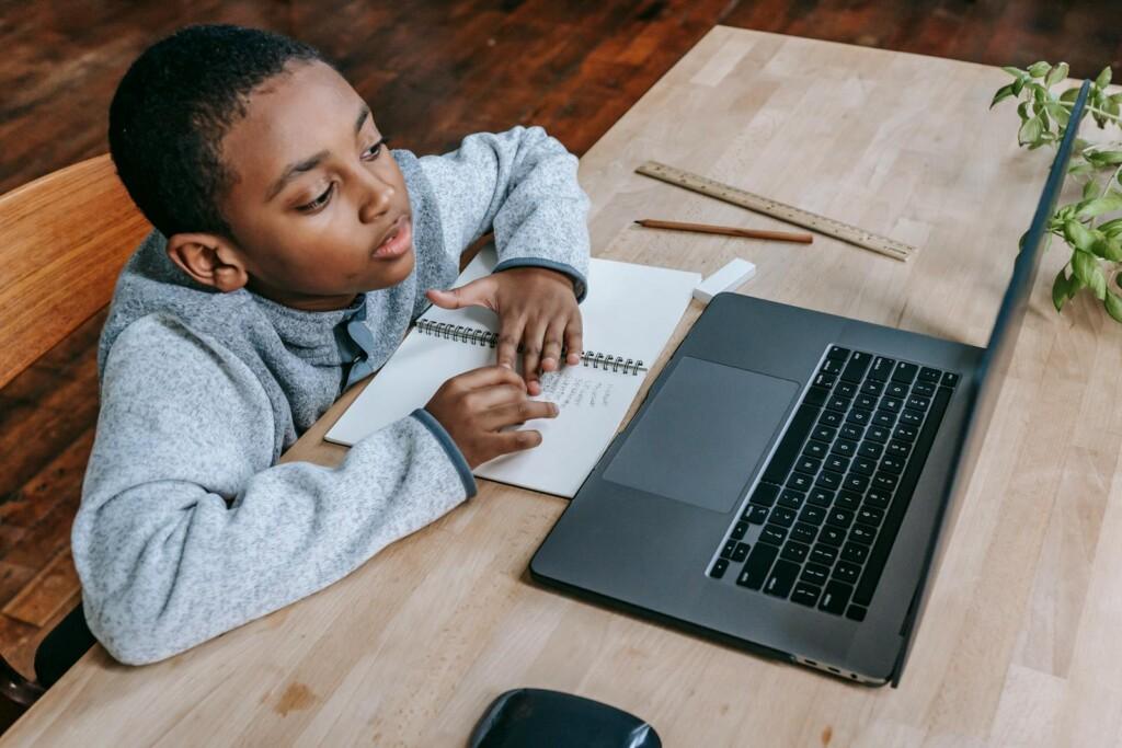 From above of smart cute African American boy studying online with netbook while sitting at table with notebook and pencil