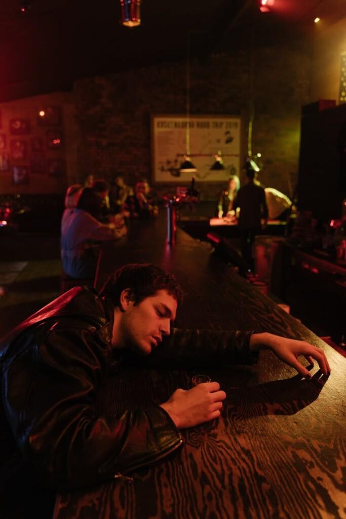 A man in a leather jacket appears sleepy at a dimly lit bar counter, late night vibe.