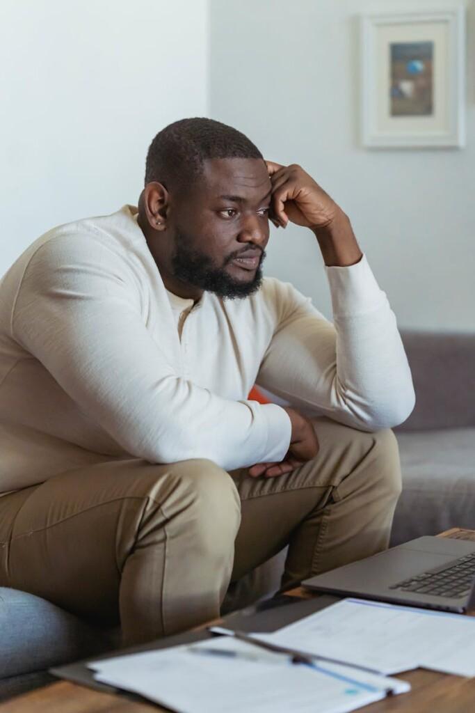 A thoughtful adult man sits in a cozy home setting, focused on his laptop.