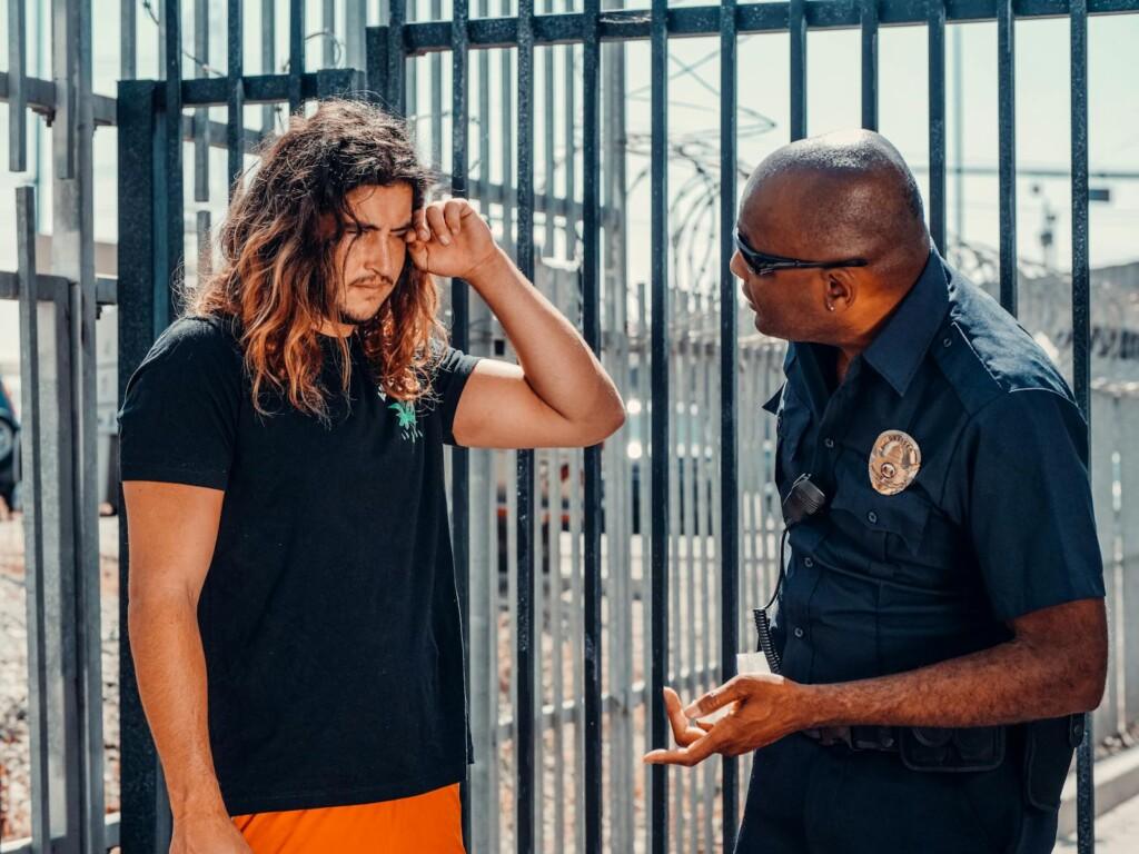 A police officer engages in a serious conversation with a civilian outdoors near a fence.