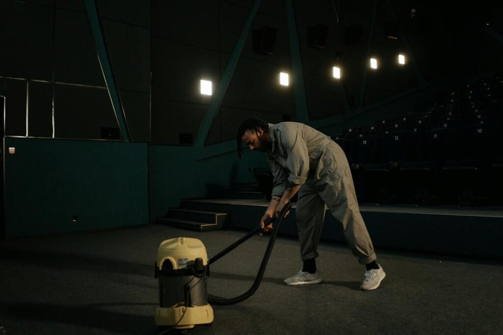 A cinema employee cleaning the theater floor with a vacuum cleaner under dim lights.