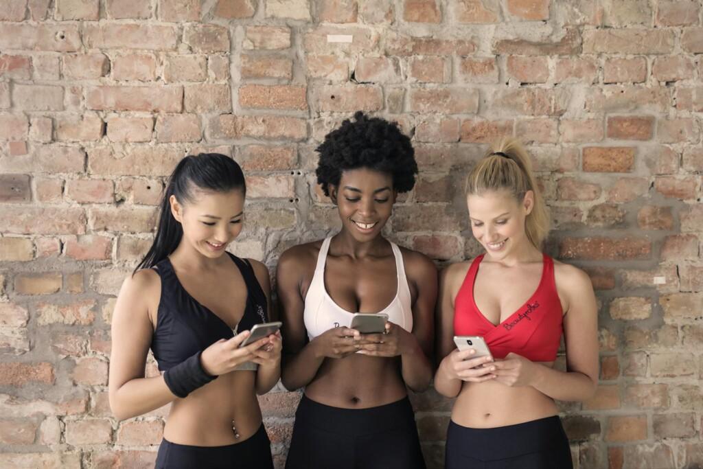 Three diverse women smiling and using smartphones indoors against a brick wall.