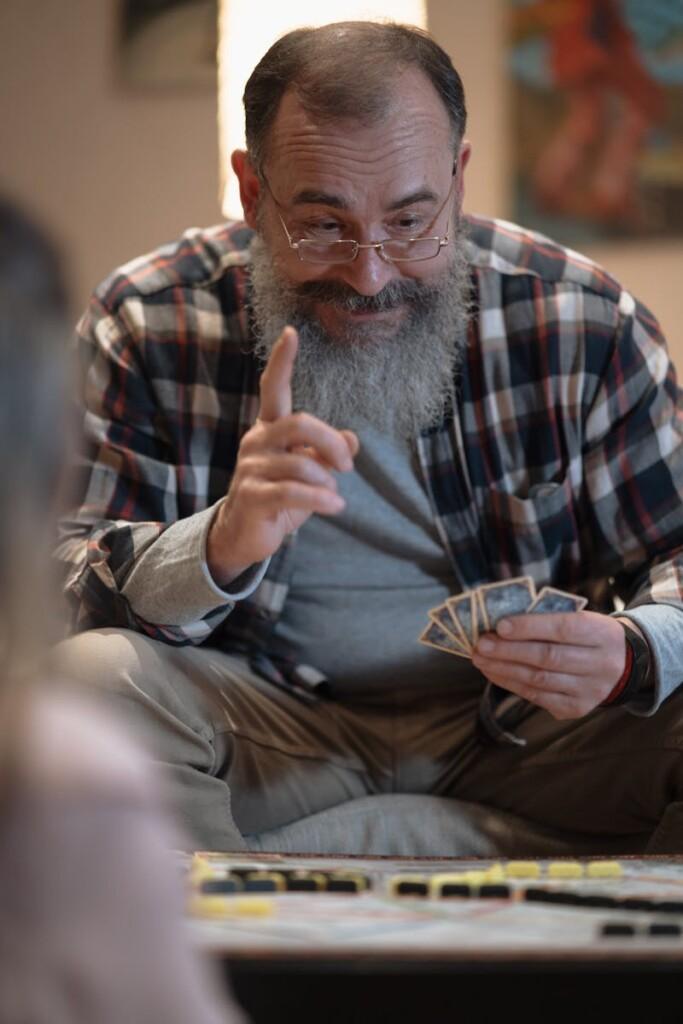 Elderly man with glasses and beard playing a board game indoors, looking focused and cheerful.